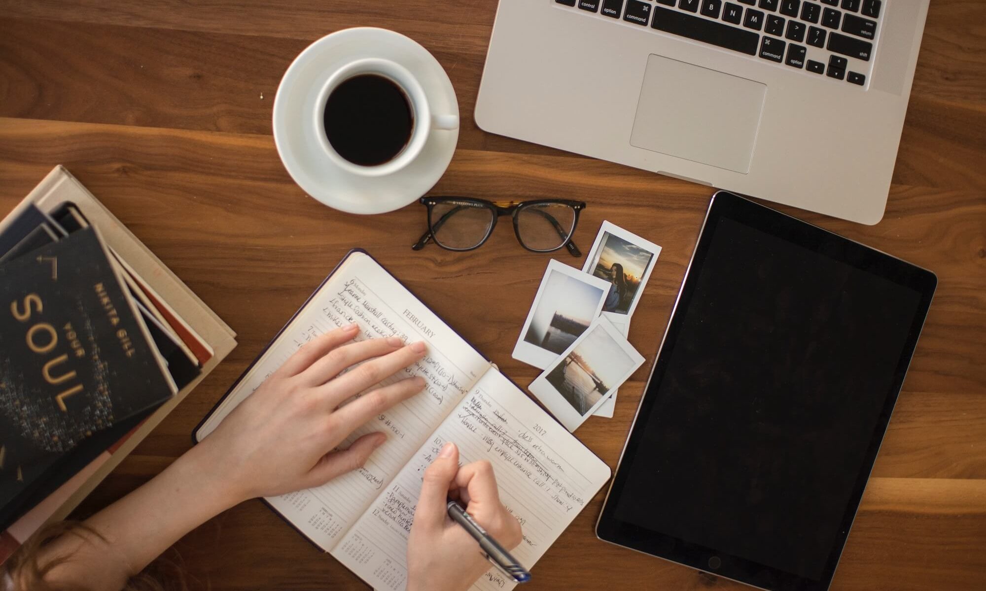 mãos escrevendo em um caderninho, com notebook e café ao lado