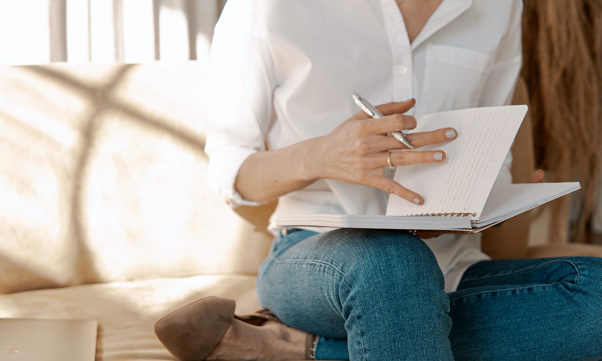 imagem de uma mulher de calça jeans e camisa branca escrevendo em um caderninho