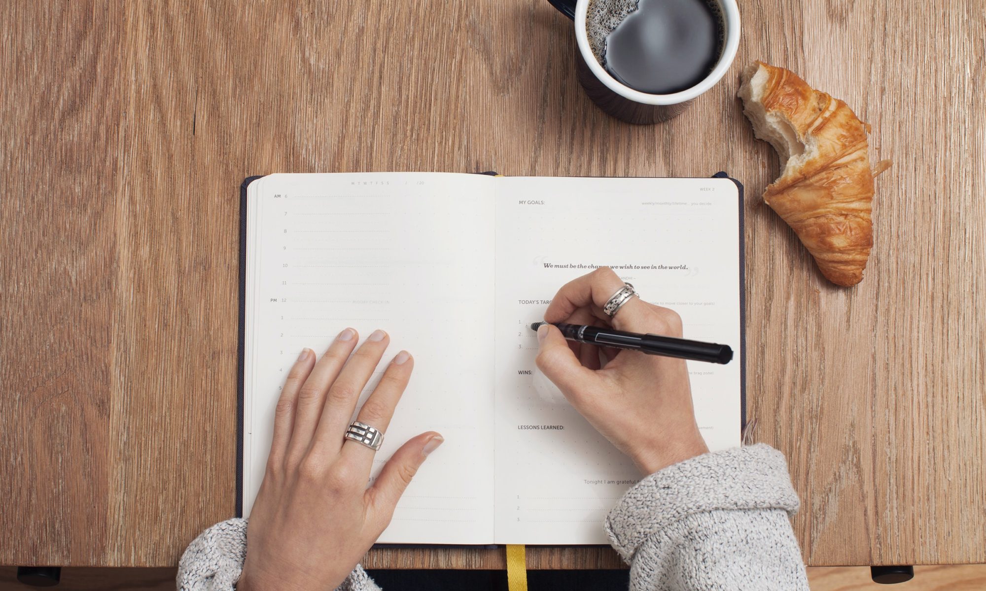 mãos escrevendo em um caderno, com uma xícara de café e croissant ao lado