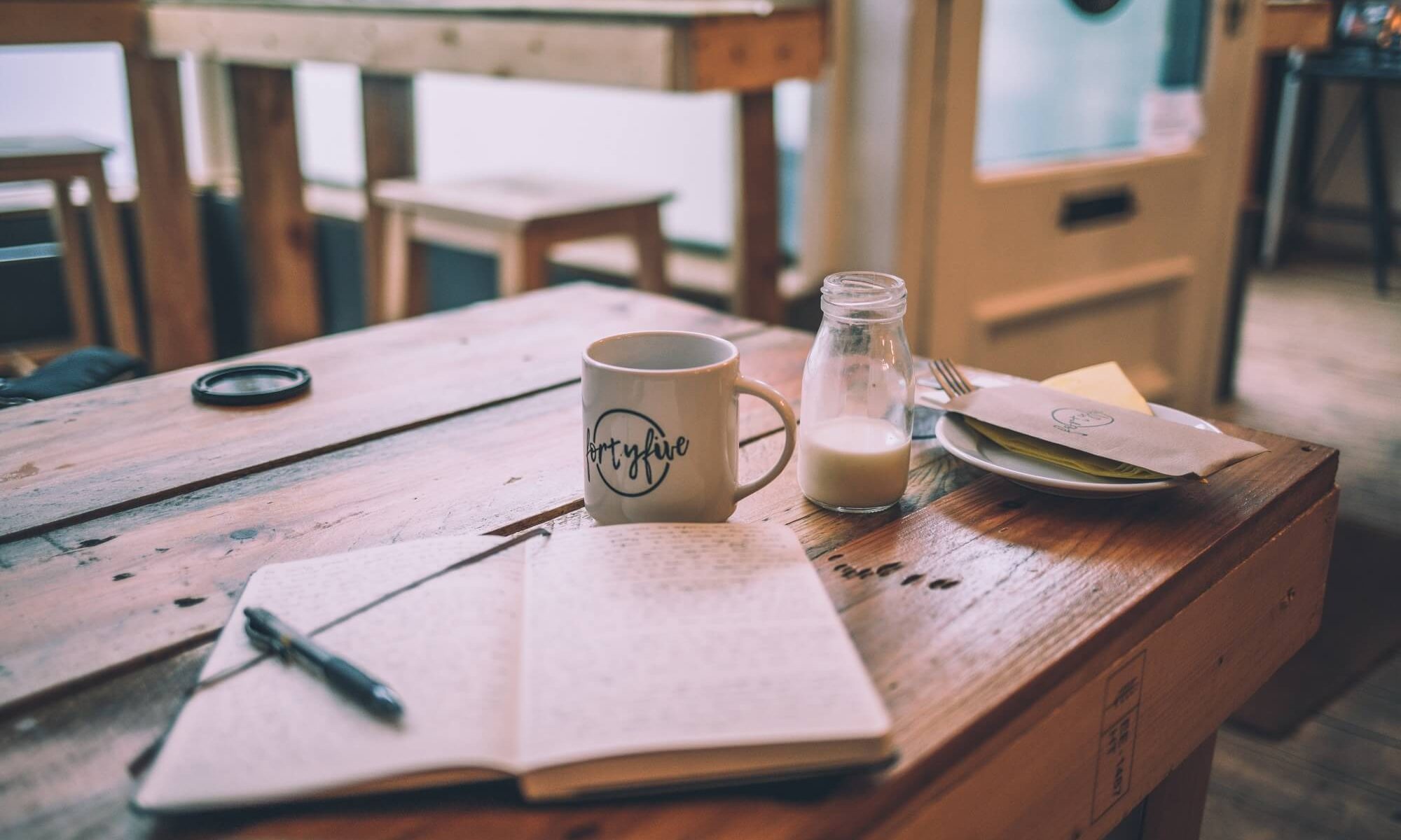 caderninho para escrita com caneta sob uma mesa de madeira com uma xícara de café e jarra de leite