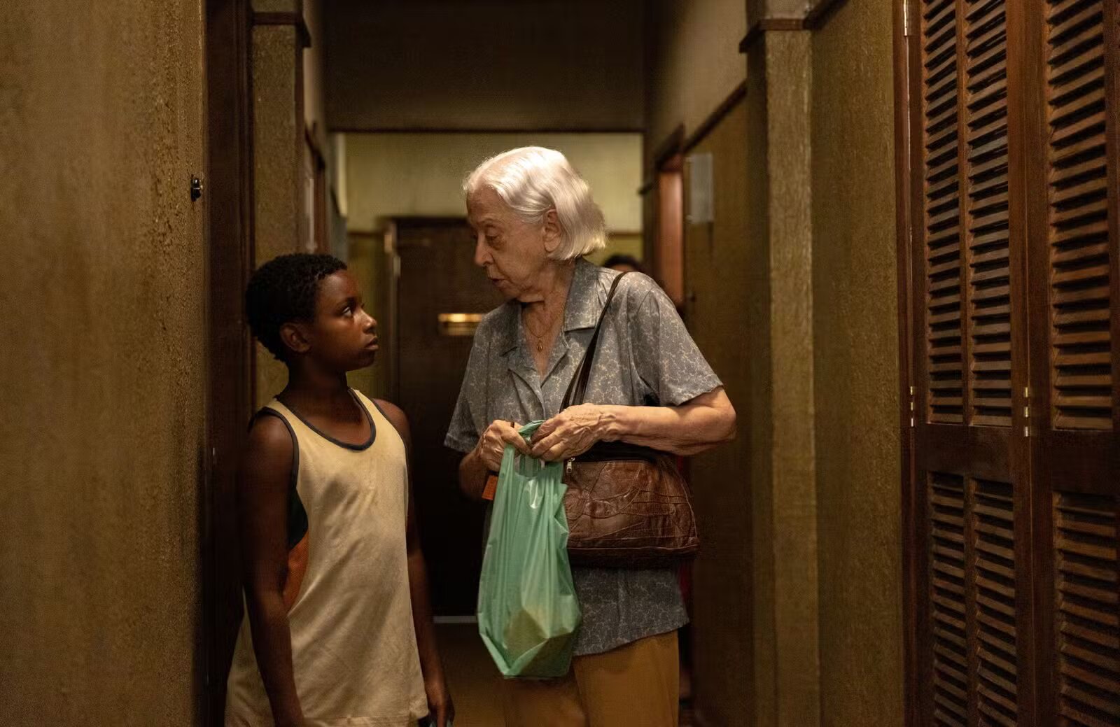 Foto de divulgação do filme "Vitória", cena entre atuantes no corredor; Dona Vitória, com uma sacola verde na mão, conversa com o jovem Marcinho;
