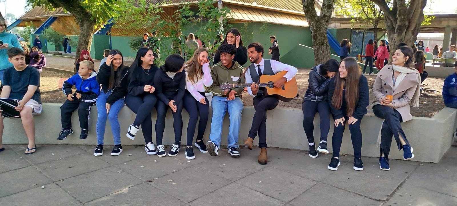 imagem mostra o músico Maurício Colina tocando canções junto às e os estudantes da EMEB Arnaldo Grin. A jornalista e escritora Rafaela Dilly Kich aparece à direita, também acompanhando as canções;